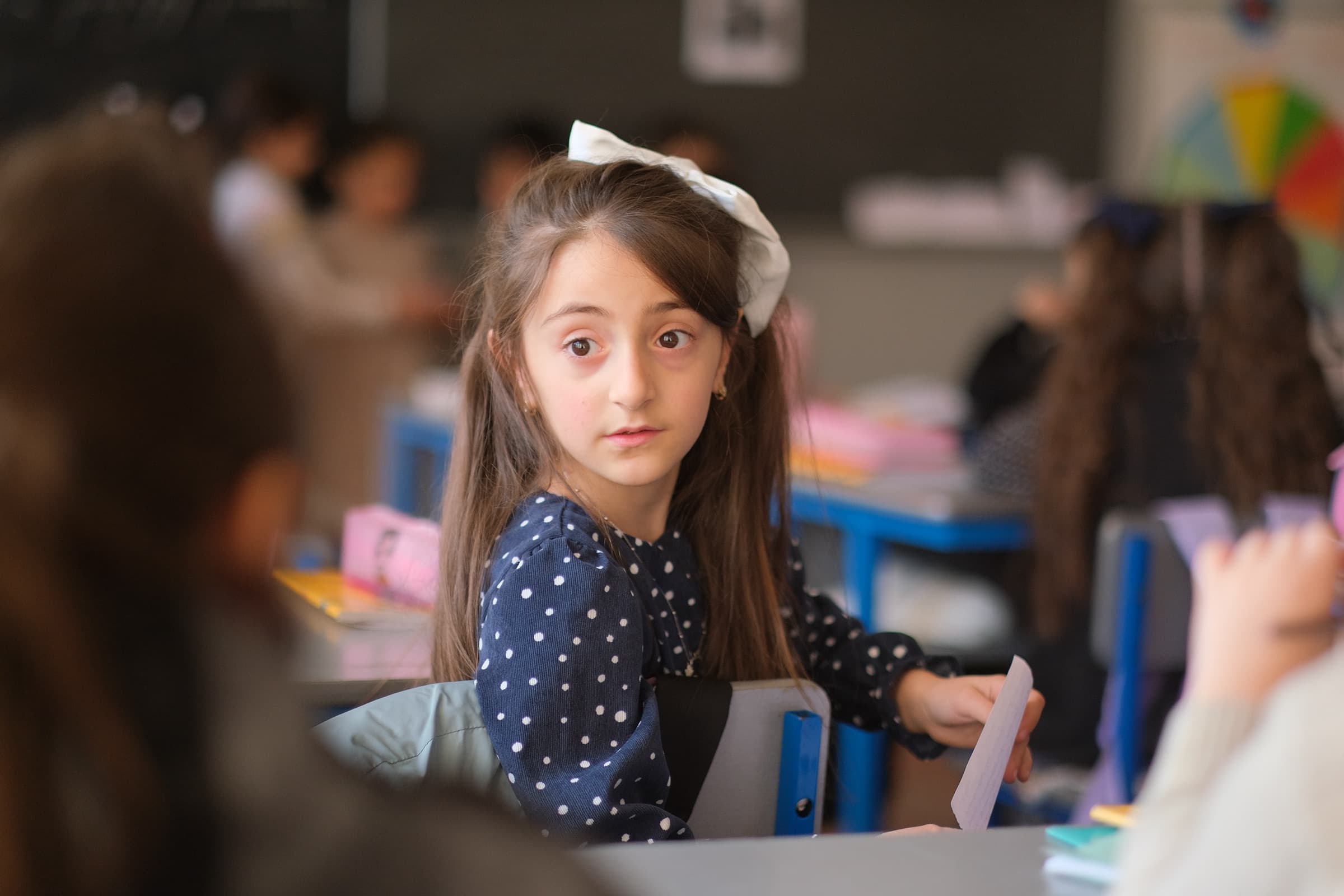 A young girl at her classroom desk looking up from her work, with other children out of focus around her